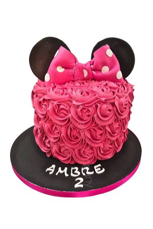 Un gâteau d'anniversaire rond entièrement recouvert de rosaces pochées en crème au beurre rose fuchsia, surmonté de deux grandes oreilles noires de Minnie Mouse et d'un nœud rose à pois blancs.