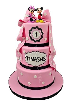 Une pièce montée d'anniversaire haut de gamme à trois étages rose, décorée de pois blancs, de rubans noirs, d'un grand nœud drapé en sucre et de figurines de Mickey et Minnie Mouse au sommet.