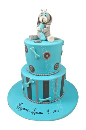 Pièce montée de 1er anniversaire à deux étages bleu turquoise et gris décorée de rayures verticales, de cercles, d'os en pâte à sucre et surmontée du doudou chien Arthur de Noukie's tenant le chiffre 1 réalisée par The French Cake Company