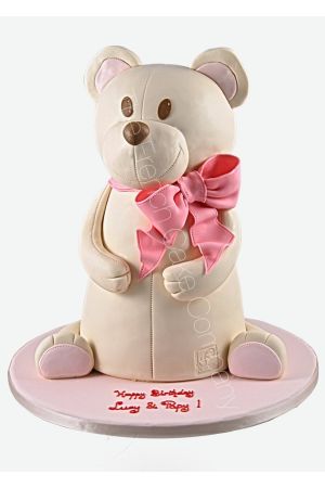 A large pastel teddy bear is placed on top of a cake, decorated with a pink bow, designed for a girl's birthday.