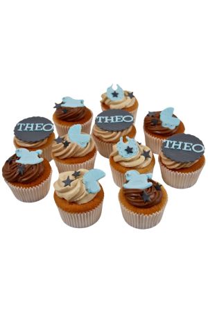 Un assortiment de cupcakes avec glaçage chocolat et crème, décorés de motifs de bébé en sucre bleus et gris tels que des landaus, des bavoirs et des chevaux à bascule.