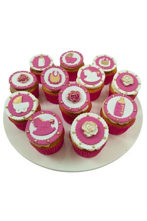 Un assortiment de cupcakes roses et blancs décorés de motifs de bébé en sucre dorés et roses, tels que des biberons, des landaus et des chevaux à bascule.