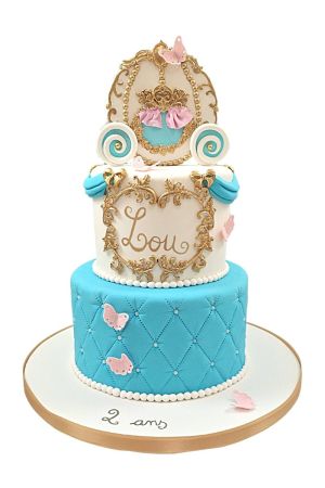 Luxe twee-laags verjaardagstaart met turquoise gewatteerde onderlaag, roze vlinders, barokke gouden krullen en een gouden Assepoester koets op de top gemaakt door The French Cake Company