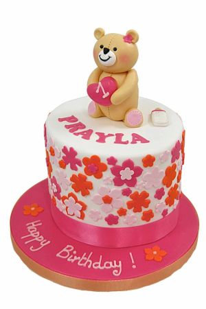 Round white 1st birthday cake decorated with pink, orange, and white fondant flowers, topped with a 3D Forever Friends teddy bear holding a pink heart with number 1 on a bright pink board by The French Cake Company
