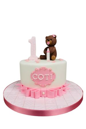 Lieve witte 1e verjaardagstaart met een bruin suikerbeertje, zuigfles, roze ruches en het getal 1 gemaakt door The French Cake Company