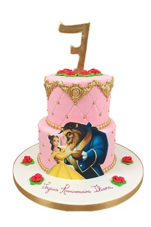 Elegant two-tier quilted pink birthday cake featuring gold baroque crowns, red fondant roses, gold pearl trim, and a printed Belle and Beast illustration cutout by The French Cake Company