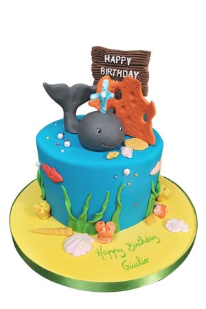 Un gâteau d'anniversaire rond bleu turquoise en pâte à sucre décoré de lianes marines et de coquillages, surmonté d'une figurine de baleine grise et d'un récif de corail orange.