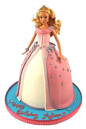 Blonde Barbie doll cake dressed in a pink and cream ballgown with lilac trim, small sugar blossoms, and textured bodice on a bright blue round cake board by The French Cake Company