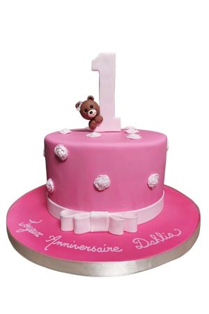Roze 1e verjaardagstaart gedecoreerd met witte suikerroosjes, een strik en een kiekeboe suikerbeertje achter het getal 1 gemaakt door The French Cake Company