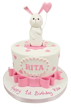 Charming white 1st birthday cake featuring a sculpted 3D bunny holding a heart balloon, pink ribbon frills, and sugar hearts by The French Cake Company