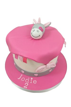 Gâteau d'anniversaire crème décoré de fanions roses, d'un ruban gris à petits cœurs et surmonté d'un doudou couverture rose vif avec tête 3D de l'âne licorne Noukie's réalisé par The French Cake Company