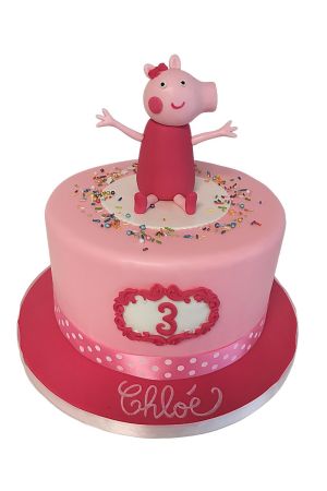 Gâteau d'anniversaire rose poudré décoré de vermicelles multicolores, d'un ruban à pois et surmonté d'une figurine 3D de Peppa Pig en robe rose fuchsia réalisée par The French Cake Company