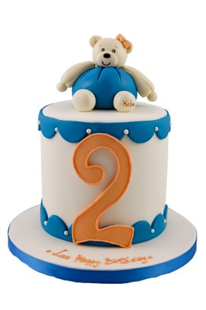 Gâteau d'anniversaire personnalisé blanc et bleu garni d'une bordure festonnée, de délicates perles, d'un grand chiffre 2 orange et d'un ourson en peluche Kaloo bleu sculpté en 3D, créé par The French Cake Company.
