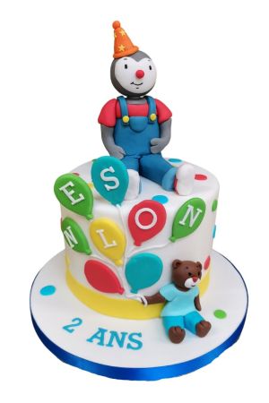 Un gâteau d'anniversaire blanc avec une figurine 3D de T'choupi, le doudou et des ballons colorés indiquant un prénom.