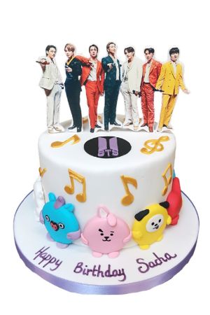 White BTS birthday cake decorated with musical notes, BT21 character figures around the base, and band member photo toppers on top