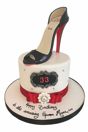 Un gâteau d'anniversaire blanc orné d'un ruban rouge et d'un médaillon baroque noir, surmonté d'un escarpin noir à talon haut comestible avec une semelle rouge emblématique.
