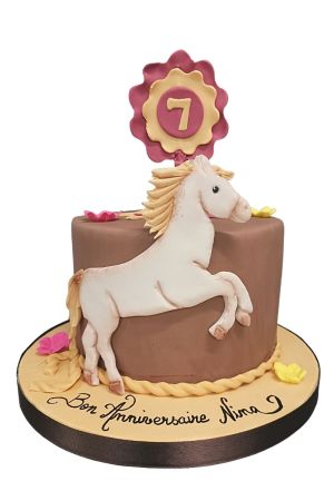 Gâteau d'anniversaire équitation marron décoré d'un cheval blanc sculpté, d'une bordure torsadée en sucre, de petites fleurs et d'un flot de compétition réalisé par The French Cake Company