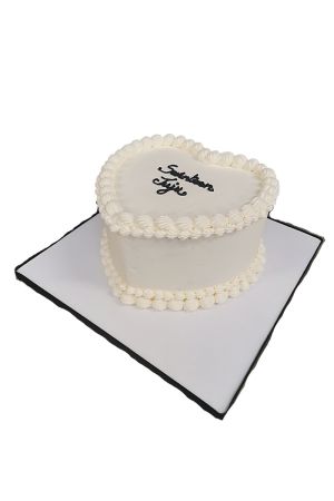 Un gâteau d'anniversaire blanc en forme de cœur, orné de bordures de crème cannelées régulières et d'un message personnalisé écrit en noir au centre.