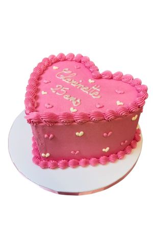 Un gâteau d'anniversaire en forme de cœur rose, décoré de bordures de crème cannelées et parsemé de petits cœurs en sucre roses et blancs.