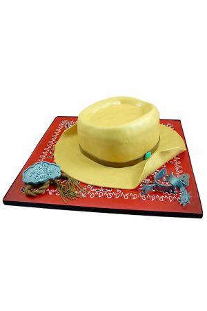 3D sculpted cream cowboy hat birthday cake with blue belt buckle, spurs, and lasso on a red bandana print base