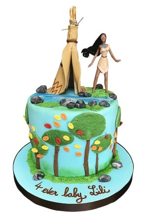 A round light blue fondant birthday cake decorated with forest trees and colorful autumn leaves, topped with a 3D tan sugar teepee tent, a flowing river with grey rocks, and a Pocahontas figurine.