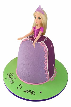 Een verjaardagstaart in de vorm van de paarse jurk van Prinses Rapunzel met een pop bovenop, gepresenteerd op een groene plaat.