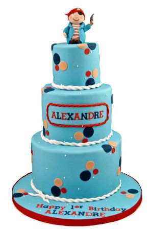A tall three-tiered light blue fondant birthday cake decorated with dark blue, orange, and red confetti dots, topped with a 3D sugar figurine of a cute smiling pirate holding a dagger.