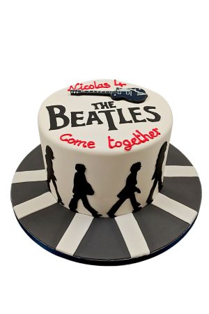 Un gâteau d'anniversaire rond en pâte à sucre blanche arborant le logo officiel des Beatles noirs et l'inscription rouge Come together, surmonté d'une guitare, posé sur un plateau reprenant le passage piéton d'Abbey Road.