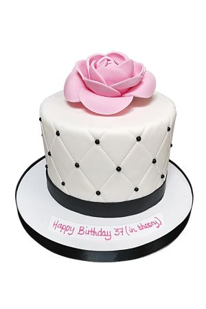 Gâteau d'anniversaire blanc matelassé orné de perles noires, d'un ruban sombre et d'une grande fleur de camélia rose en sucre réalisé par The French Cake Company
