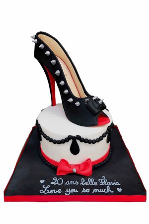 Un gâteau d'anniversaire blanc décoré de perles noires et d'un nœud rouge, surmonté d'une chaussure à talon noire comestible avec des clous argentés.