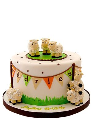 Gâteau de baptême blanc à un étage décoré de petits moutons en sucre en 3D, d'herbe verte et d'une guirlande de fanions.