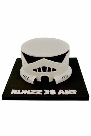 Un gâteau d'anniversaire en forme de casque de Stormtrooper blanc, noir et gris avec des détails complexes, posé sur un plateau noir.