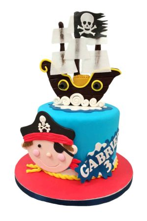A round blue fondant birthday cake decorated with a large flat-lay cartoon pirate face wearing a skull hat, topped with a detailed 3D brown pirate ship sailing on sculpted white waves with a black skull and crossbones flag.