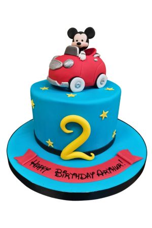 Gâteau d'anniversaire bleu vif orné d'étoiles jaunes et du chiffre 2, surmonté d'une figurine 3D en sucre de Mickey Mouse au volant d'une voiture rouge, créé par The French Cake Company.
