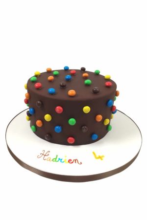 Un gâteau d'anniversaire rond en pâte à sucre marron, décoré de bonbons M&M's multicolores disposés sur le dessus et les côtés.