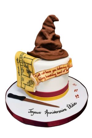 Un gâteau d'anniversaire rond en pâte à sucre blanche ceinturé de rubans bordeaux et jaune, surmonté d'un grand Choixpeau magique marron en 3D, d'un rouleau comestible représentant la Carte du Maraudeur et d'une baguette.