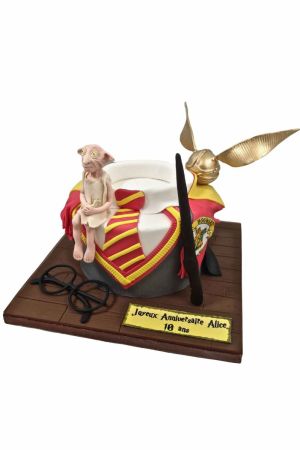 Un gâteau d'anniversaire rond en pâte à sucre sculpté en forme d'uniforme de Poudlard avec cravate rouge et jaune, surmonté d'un elfe Dobby en sucre, d'un Vif d'Or doré, d'une baguette et de lunettes noires.