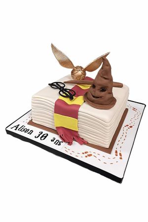 Un gâteau d'anniversaire carré en pâte à sucre sculpté en forme de livre de magie blanc, orné d'une écharpe rouge et jaune, surmonté d'un Choixpeau marron en 3D, d'un Vif d'Or, d'une baguette et de lunettes.