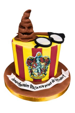 Un gâteau d'anniversaire rond en pâte à sucre aux rayures jaunes et bordeaux de Gryffondor avec son blason, surmonté d'un grand Choixpeau magique marron modelé en 3D, de lunettes noires et d'une baguette.