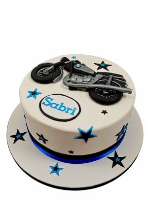 Un gâteau d'anniversaire blanc rond décoré d'étoiles noires et bleues, d'un macaron personnalisé et d'une moto BMW noire et argentée en sucre au sommet.
