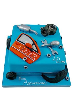 Un gâteau d'anniversaire carré bleu pétrole décoré de pièces automobiles en sucre, incluant des pneus noirs, des clés métalliques, des écrous et une portière d'auto orange.