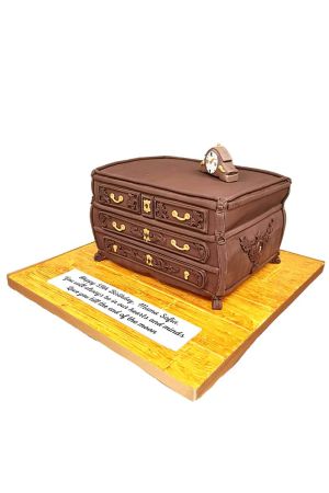 Un gâteau d'anniversaire de luxe sculpté en 3D sous la forme d'une commode en bois ancien marron avec des poignées dorées et une horloge rétro au sommet.