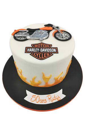 Un gâteau d'anniversaire blanc décoré de flammes en sucre orange et jaunes, du logo officiel Harley-Davidson et d'une moto en sucre orange et argentée au sommet.