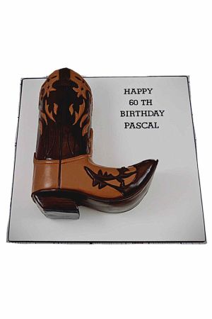 Un gâteau d'anniversaire sculpté en 3D représentant fidèlement une botte de cowboy marron et beige avec des motifs gravés, sur un plateau blanc.