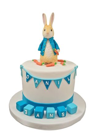 Gâteau d'anniversaire blanc élégant décoré de guirlandes de fanions bleus, de petites carottes en sucre, de cubes d'éveil au pied et surmonté d'une figurine 3D de Pierre Lapin en veste bleue réalisé par The French Cake Company