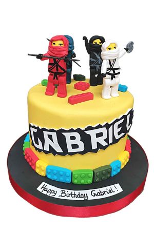Gâteau d'anniversaire personnalisé Lego Ninjago jaune, bordé de briques de construction multicolores en sucre et surmonté de figurines ninjas 3D sculptées à la main, créé par The French Cake Company.