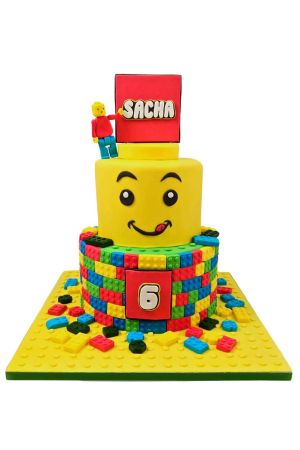 Gâteau d'anniversaire personnalisé à deux étages Lego avec base en briques multicolores, étage supérieur en forme de tête de figurine jaune souriante et personnage 3D au sommet, créé par The French Cake Company.