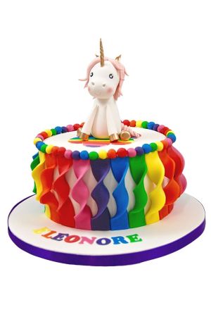 Custom round birthday cake with ruffled vertical rainbow fondant ribbons around the sides, colorful sugar pearls along the rim, and a sitting 3D unicorn figure with a gold horn, by The French Cake Company.