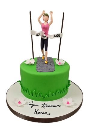 Un gâteau d'anniversaire rond vert en pâte à sucre représentant un jardin avec un chemin en pierre, surmonté d'une figurine de femme qui court et franchit une ligne d'arrivée.