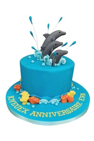 Un gâteau d'anniversaire rond bleu turquoise en pâte à sucre, surmonté de trois dauphins gris modelés jaillissant de vagues blanches et d'éclats d'eau, entouré de coraux au bas.